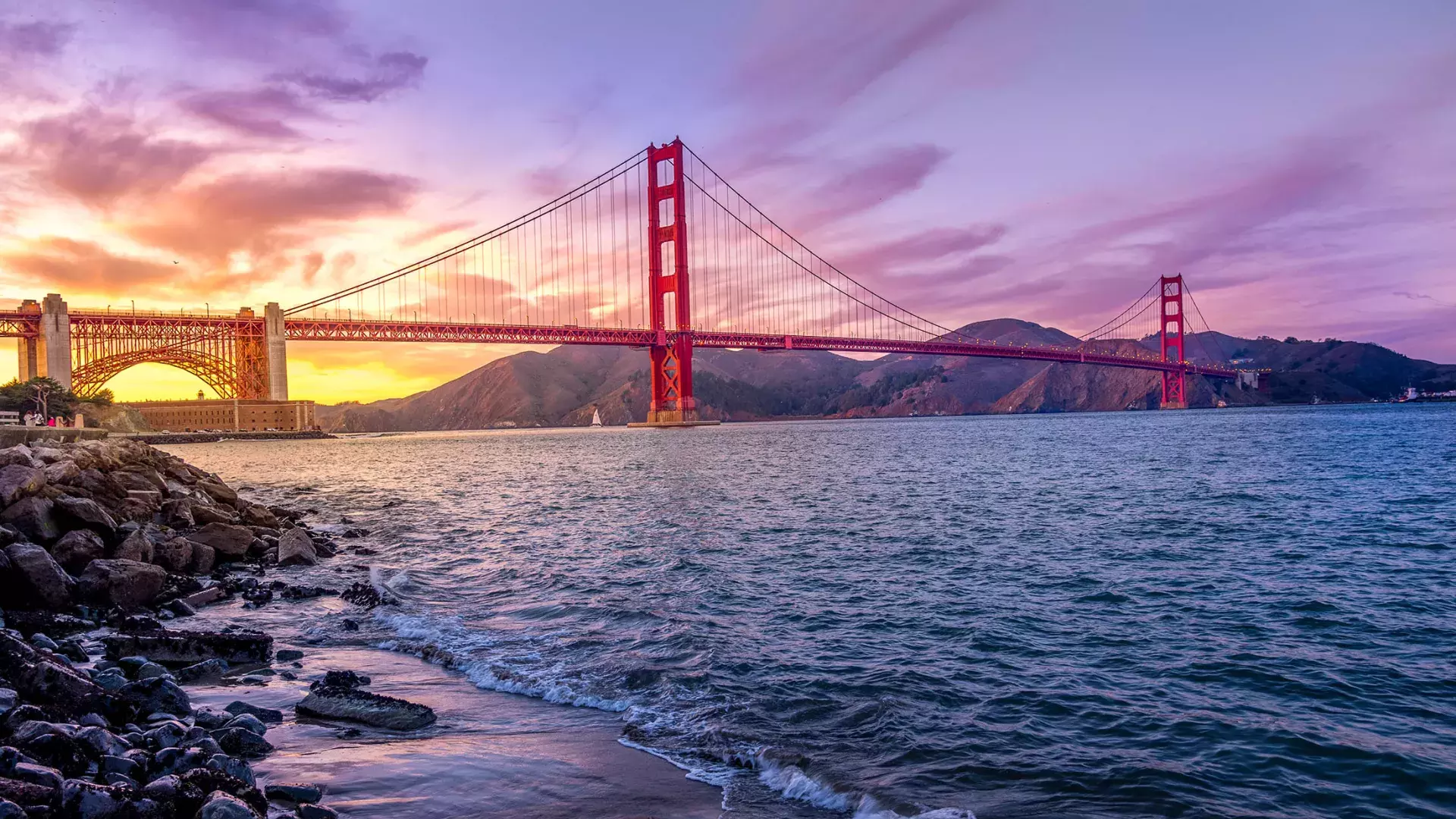 A Ponte Golden Gate ao pôr do sol com um céu multicolorido e a Baía de São Francisco em primeiro plano.