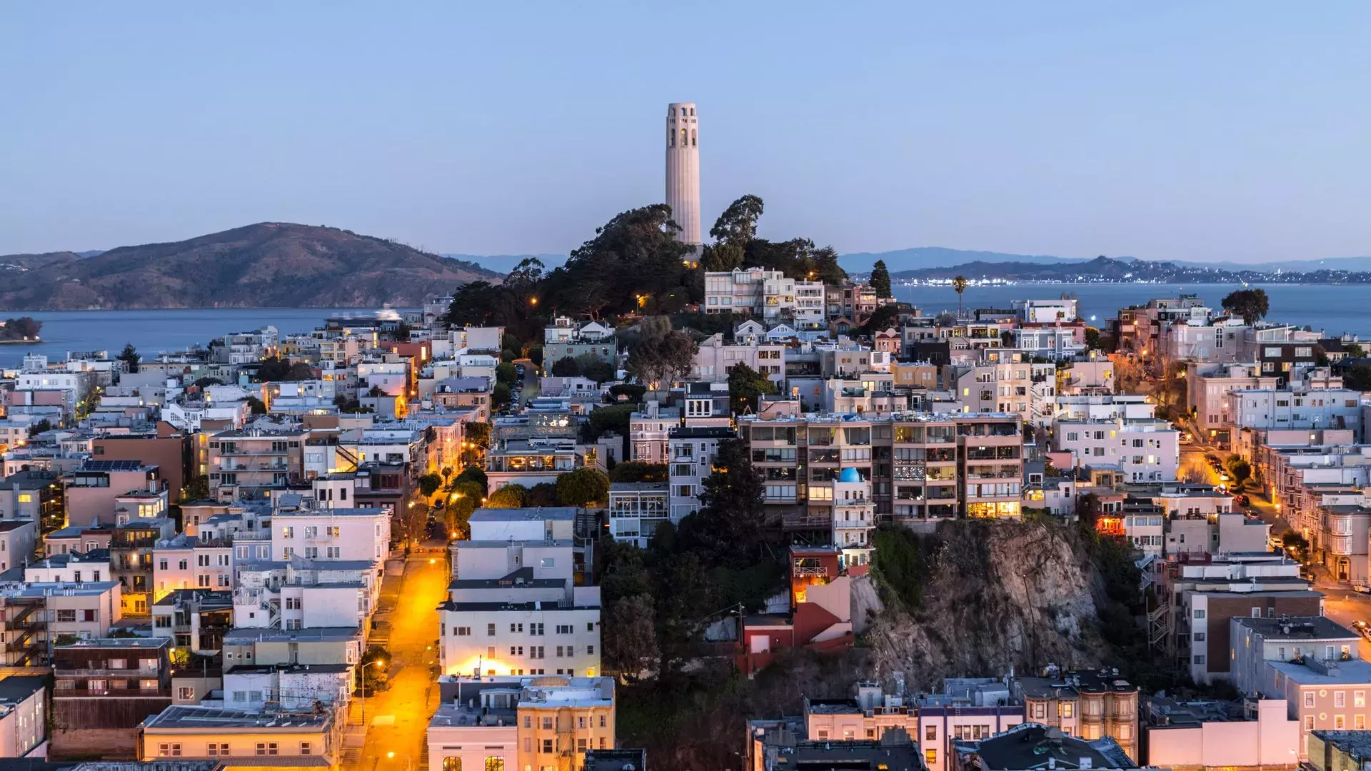 Coit Tower de São Francisco ao entardecer, com ruas iluminadas à frente e a Baía de São Francisco atrás.