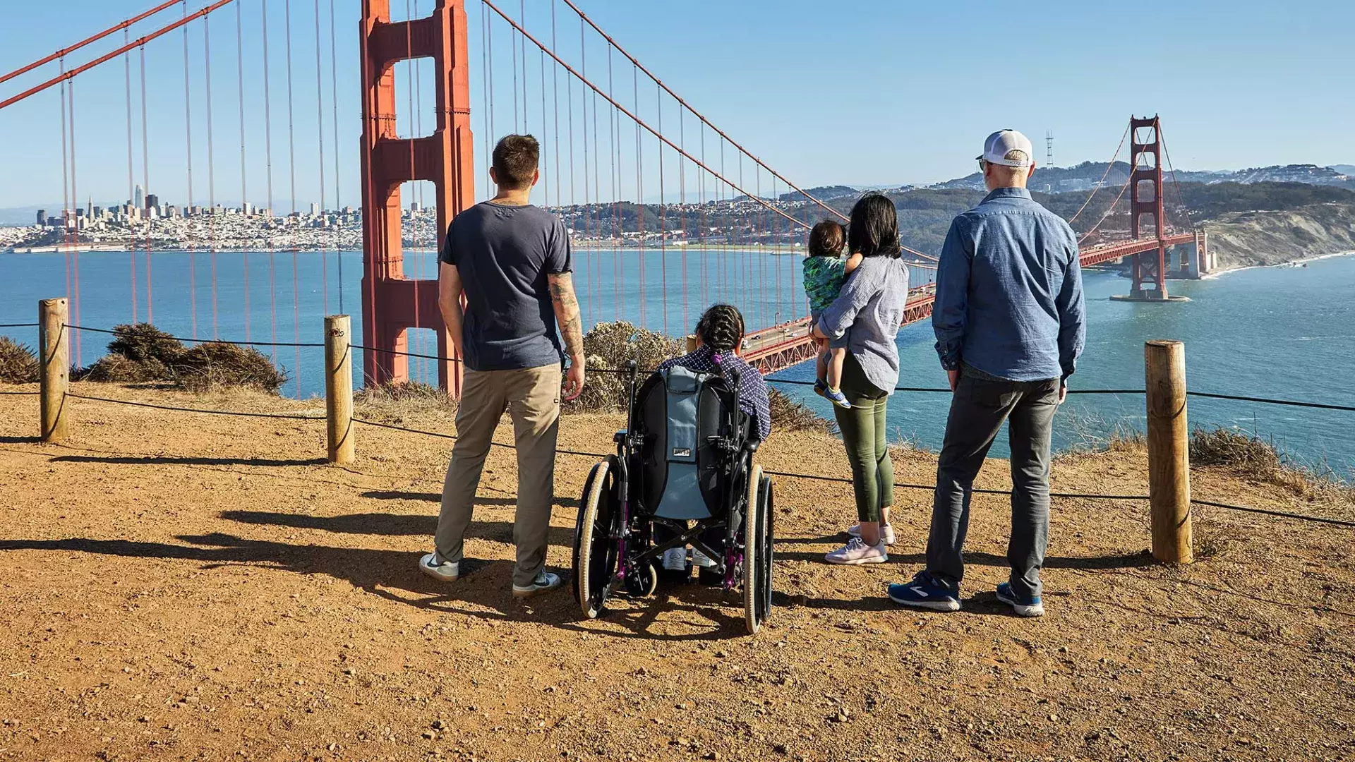 Um grupo de pessoas, incluindo uma pessoa em cadeira de rodas, é visto por trás enquanto olham para a Ponte Golden Gate de Marin Headlands.