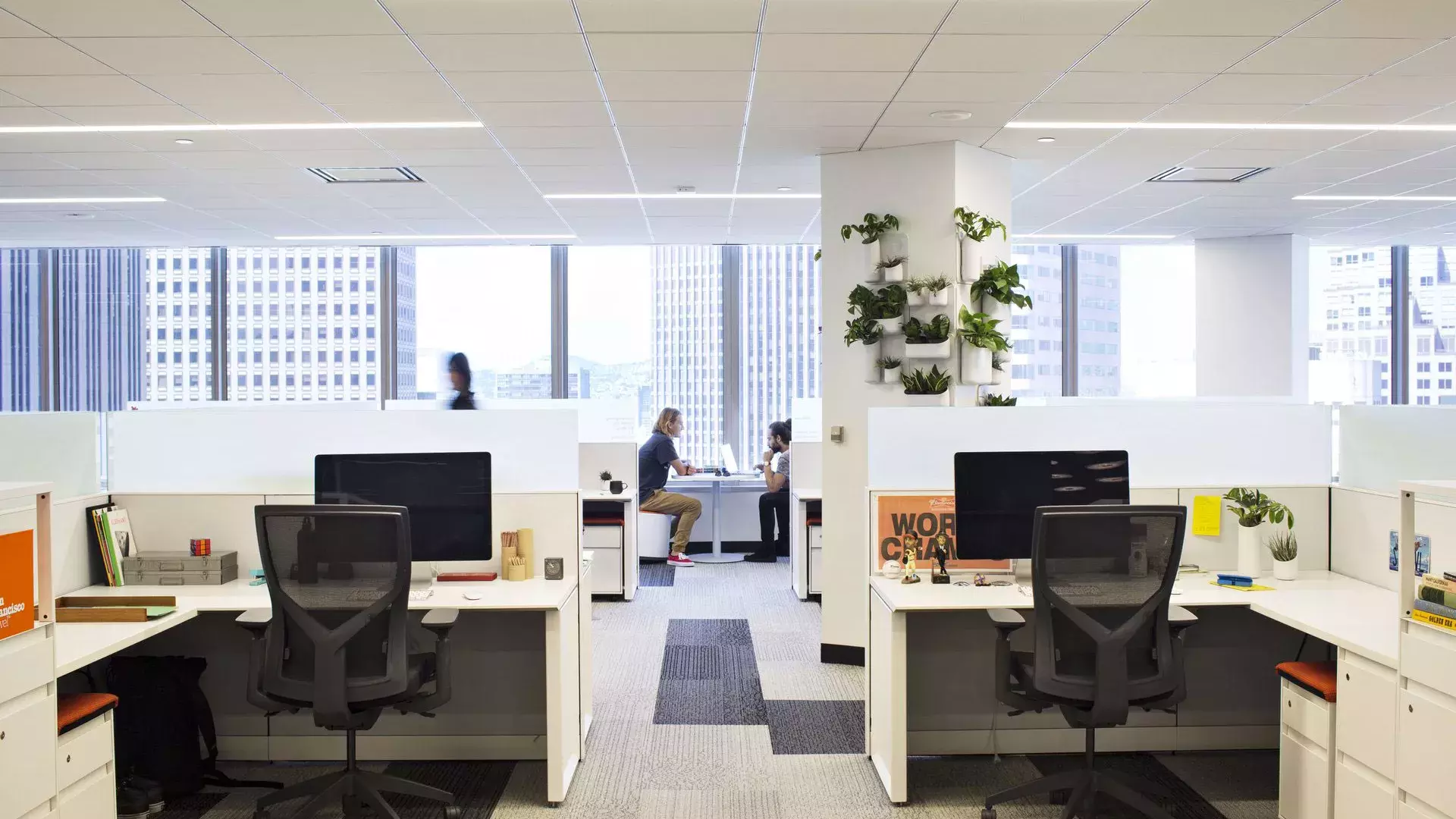 Interior scene at an office building in San Francisco, with desks in the foreground and two people working at a table in front of large windows.