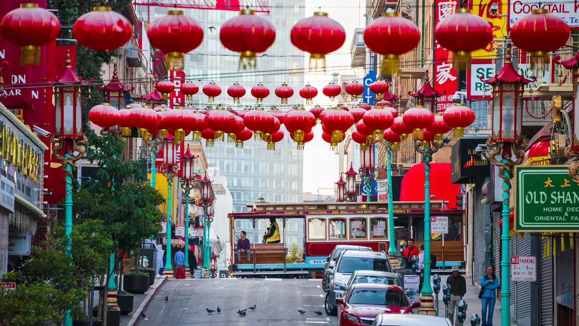 Uma rua montanhosa na Chinatown de São Francisco é retratada com lanternas vermelhas penduradas e um bonde passando.