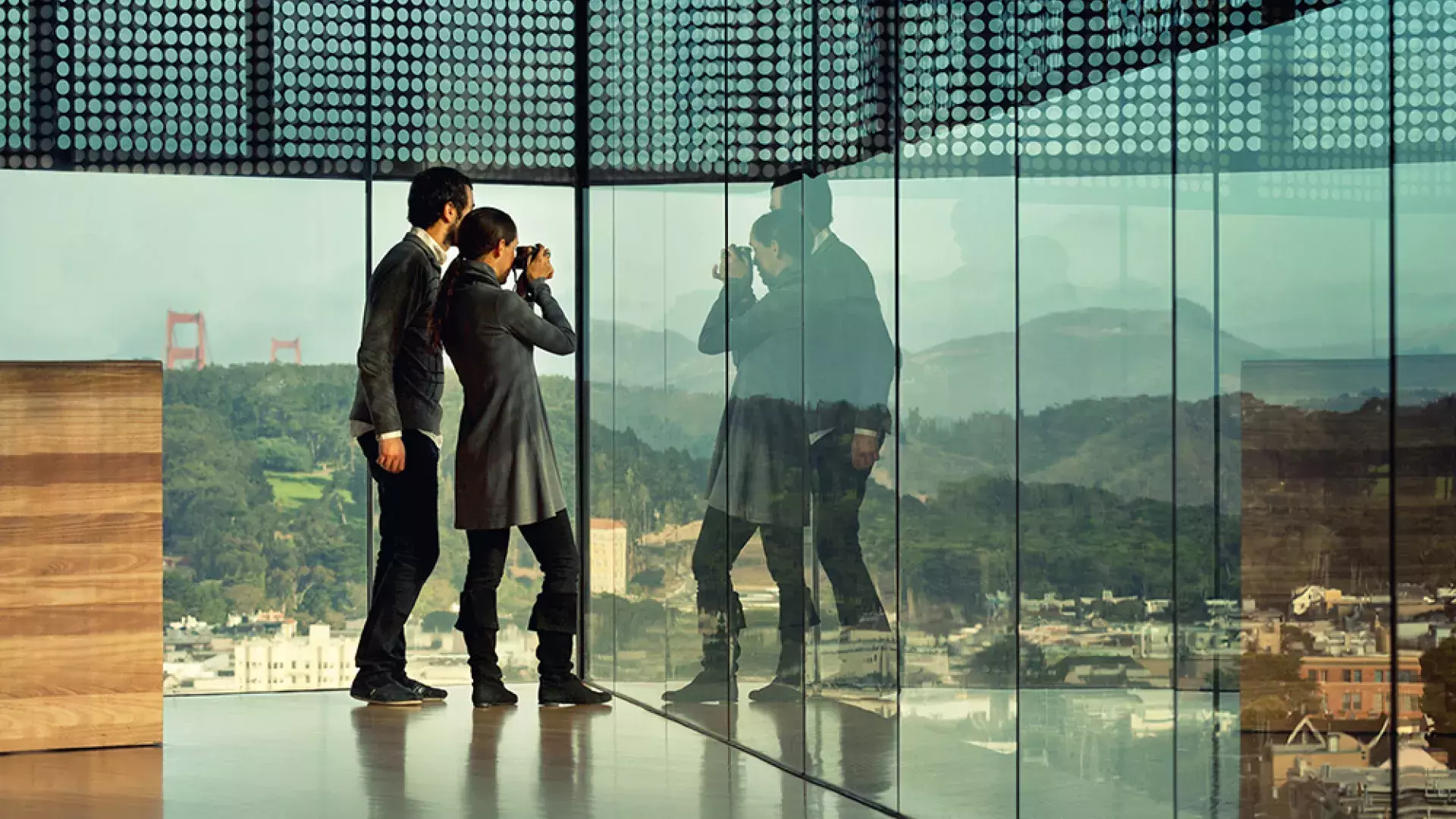 Two people peer through glass walls at the de Young Museum in San Francisco.