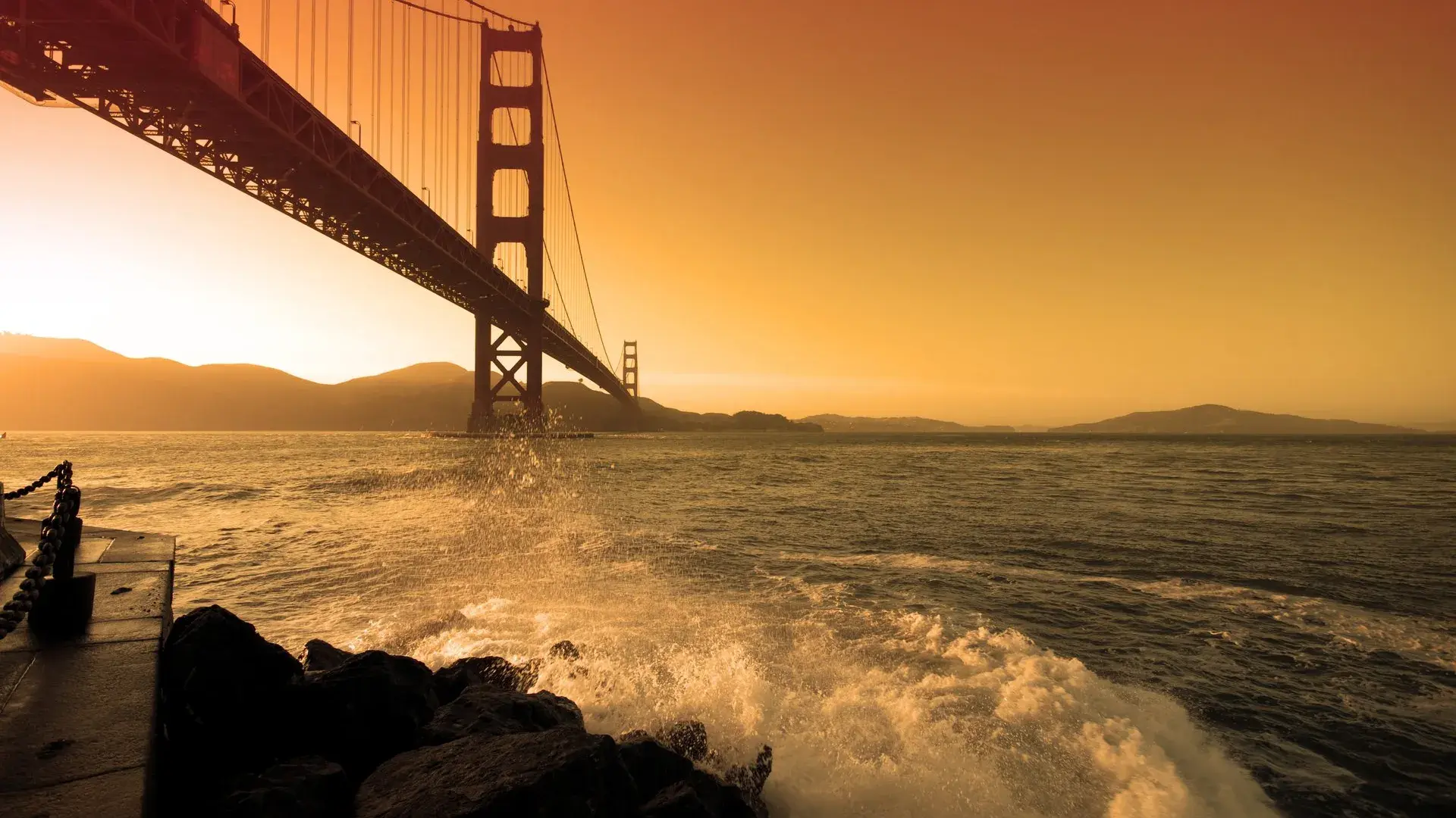 Waves crash near Fort Point beneath the Golden Gate Bridge at sunset.