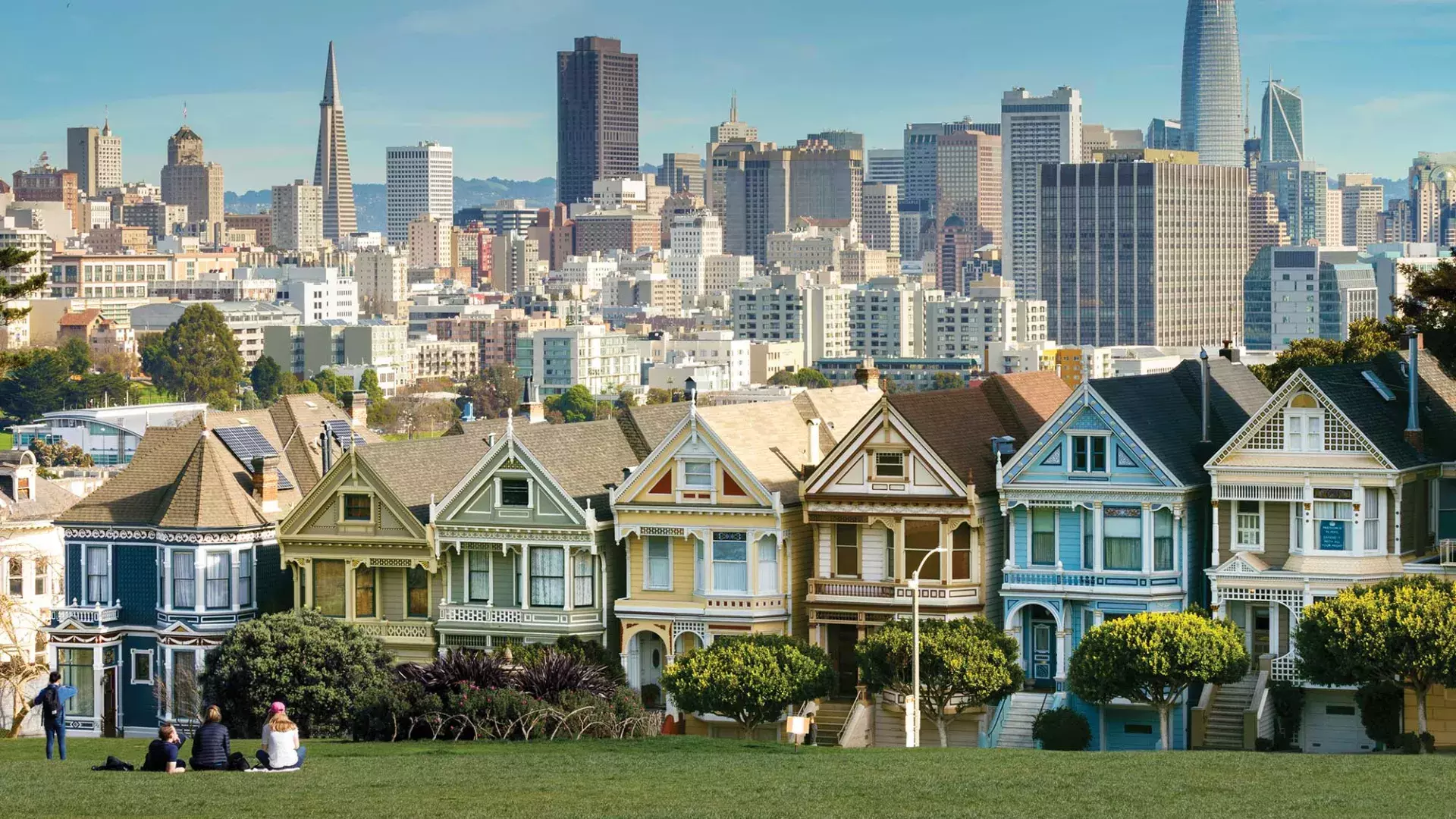 Picnickers sit on the grass at Alamo Square Park with the Painted Ladies and San Francisco skyline in the background.