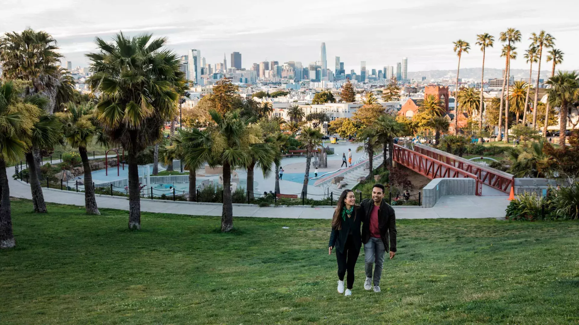 A couple walks toward the camera with Dolores Park and the San Francisco Skyline behind them.