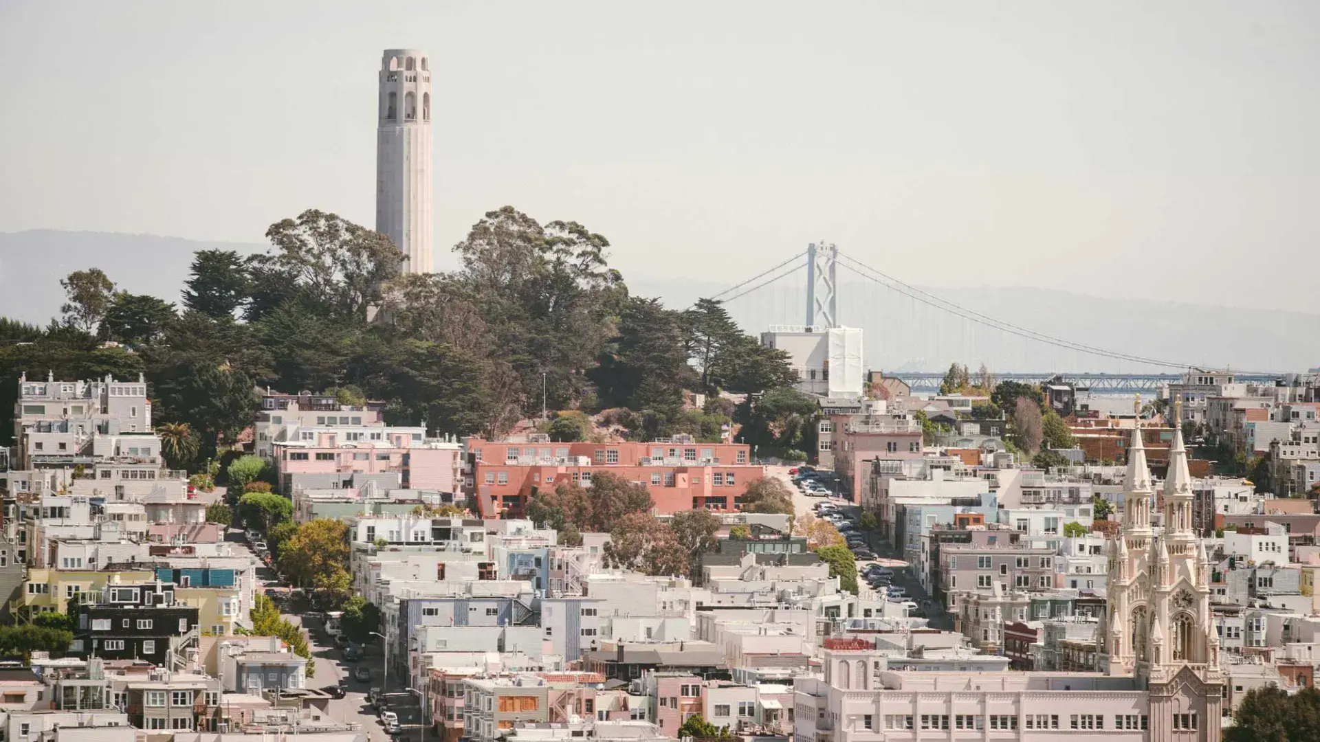 A Coit Tower de São Francisco é retratada com a Bay Bridge ao fundo e uma colina coberta de casas em primeiro plano.