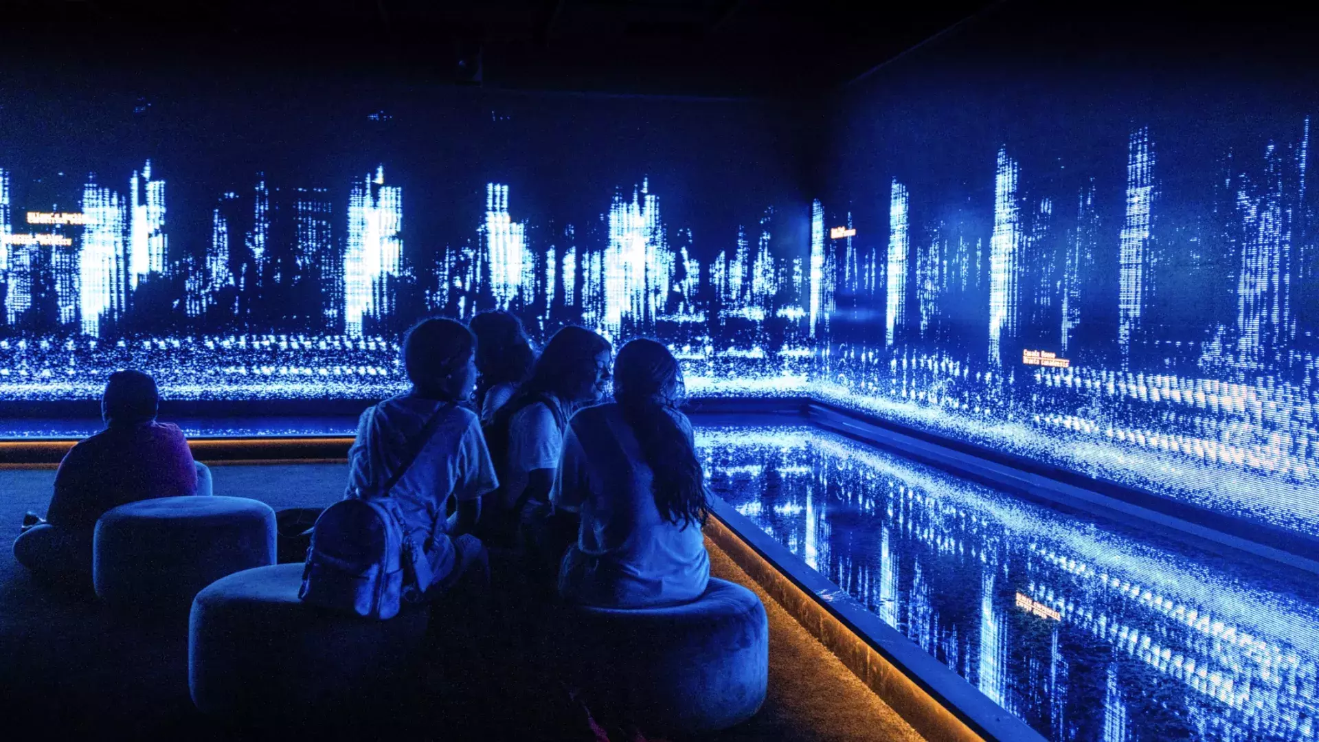 Children sit in a dark room, and look at an art installation creative of tiny moving lights on a wall