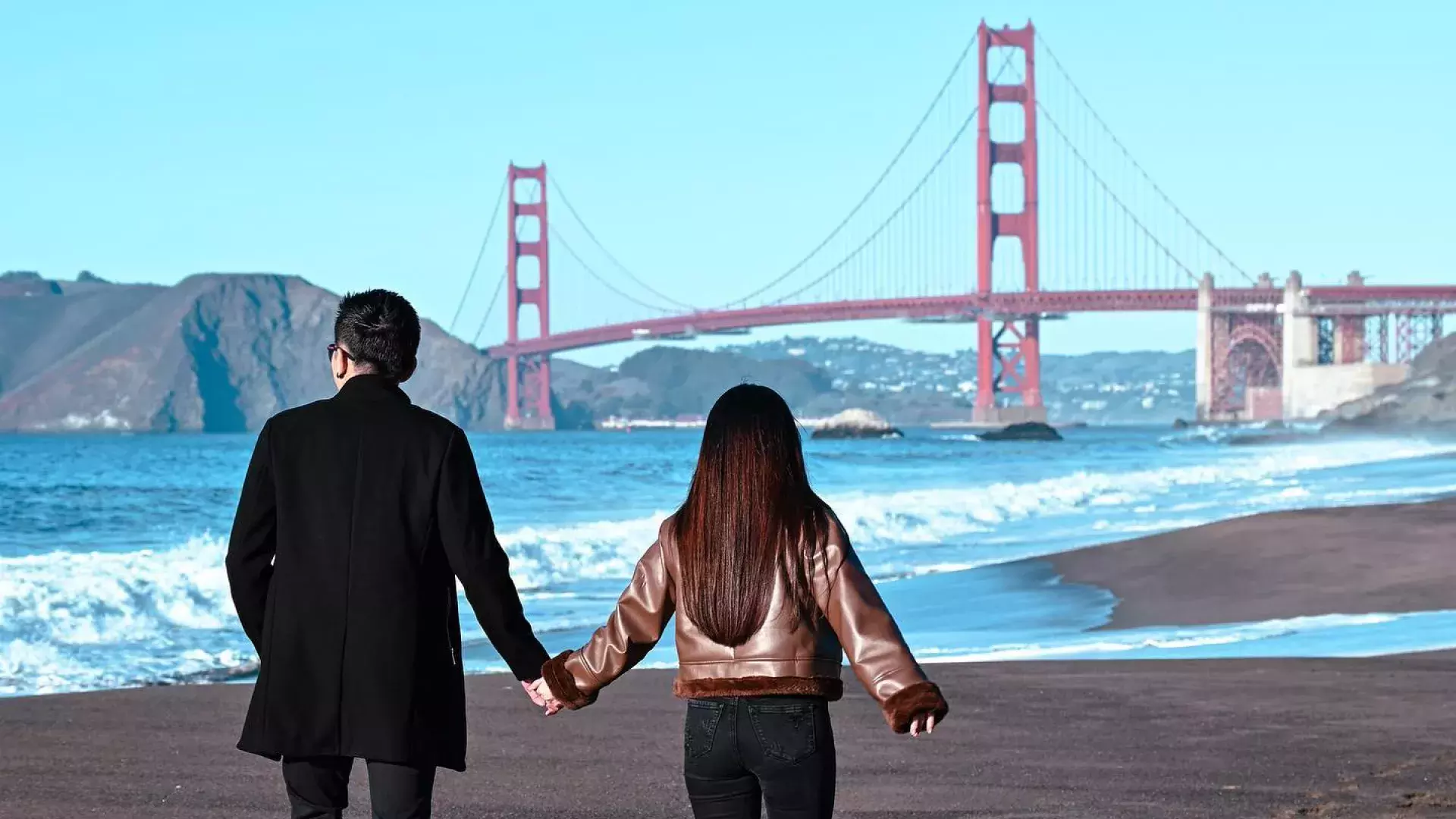 Couple holding hands at Baker Beach with Golden Gate bridge in background 