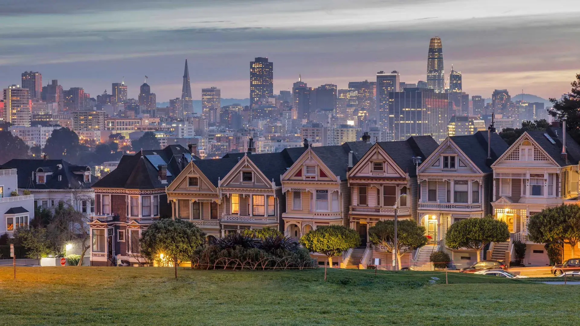 San Francisco's Painted Ladies, a row of Victorian houses, are pictured against a backdrop of the city skyline at twilight.