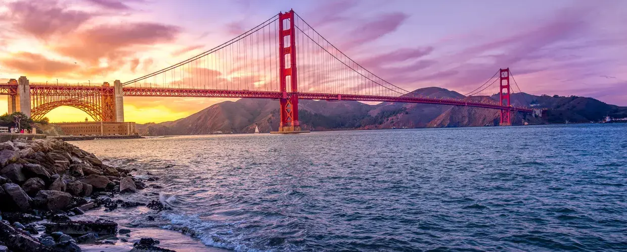 The Golden Gate Bridge at sunset with a multicolored sky and the San Francisco Bay in the foreground.
