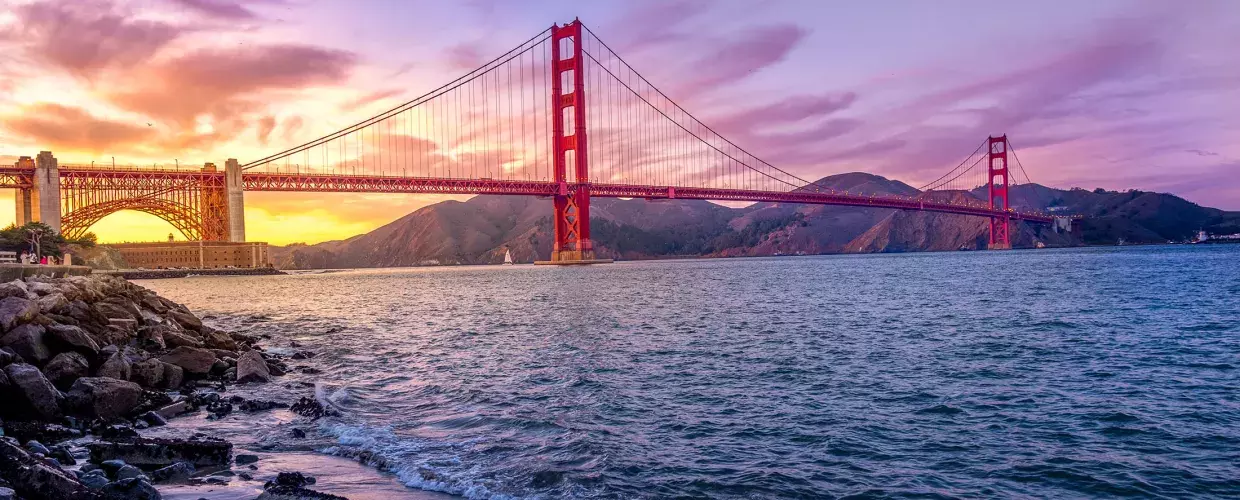 A Ponte Golden Gate ao pôr do sol com um céu multicolorido e a Baía de São Francisco em primeiro plano.