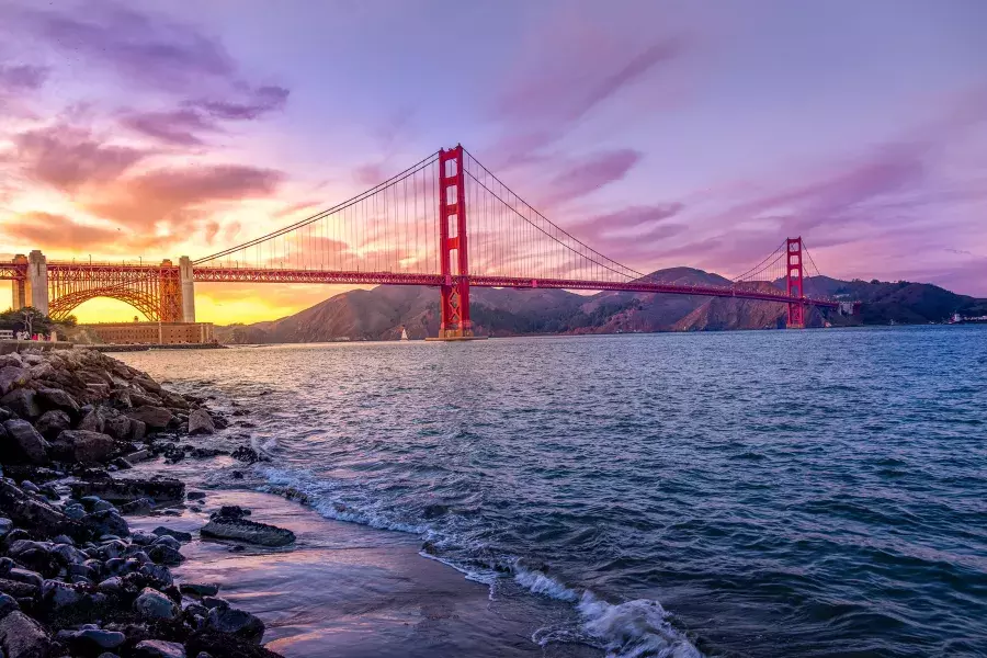 A Ponte Golden Gate ao pôr do sol com um céu multicolorido e a Baía de São Francisco em primeiro plano.