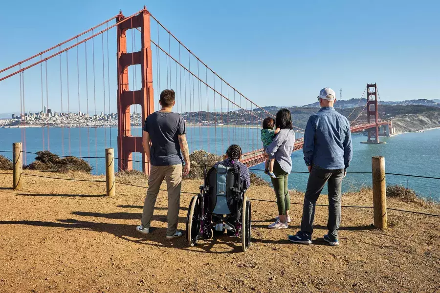 A group of people, including one person in a wheelchair, is seen from behind as they look at the Golden Gate Bridge from the Marin Headlands.