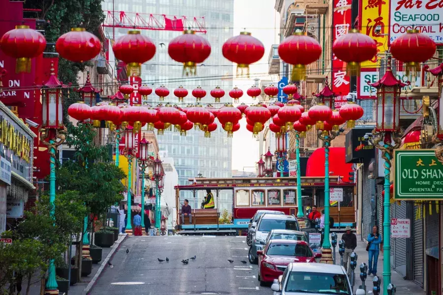 Uma rua montanhosa na Chinatown de São Francisco é retratada com lanternas vermelhas penduradas e um bonde passando.