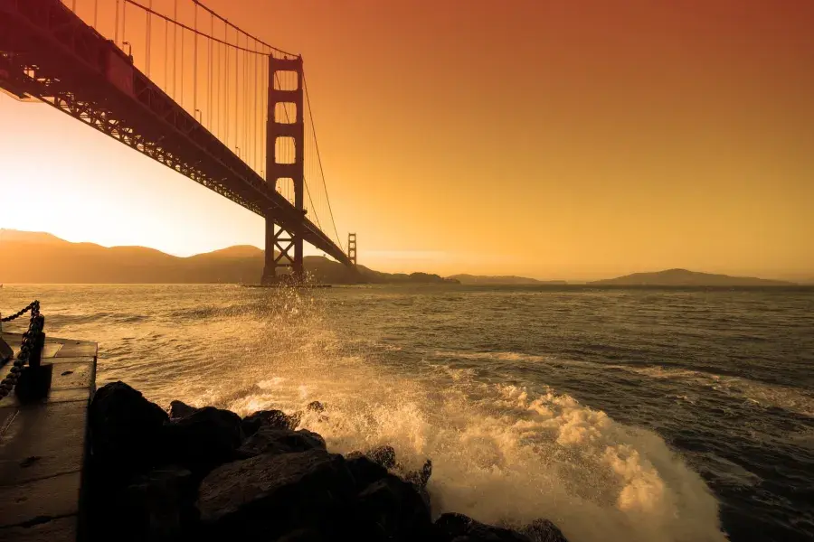 Waves crash near Fort Point beneath the Golden Gate Bridge at sunset.