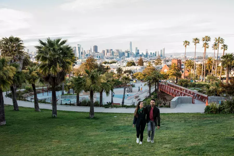 A couple walks toward the camera with Dolores Park and the San Francisco Skyline behind them.