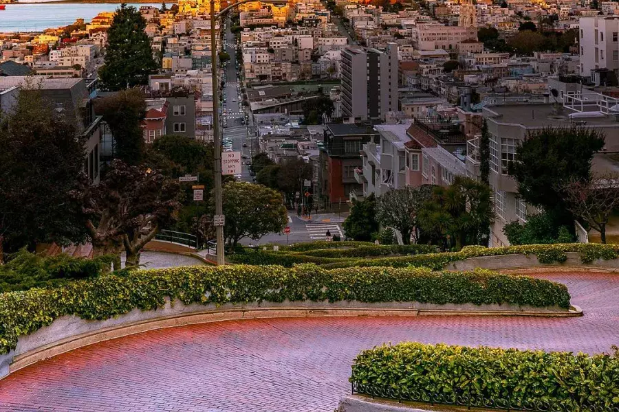 Lombard Street curves with Coit Tower in the distance during sunset.