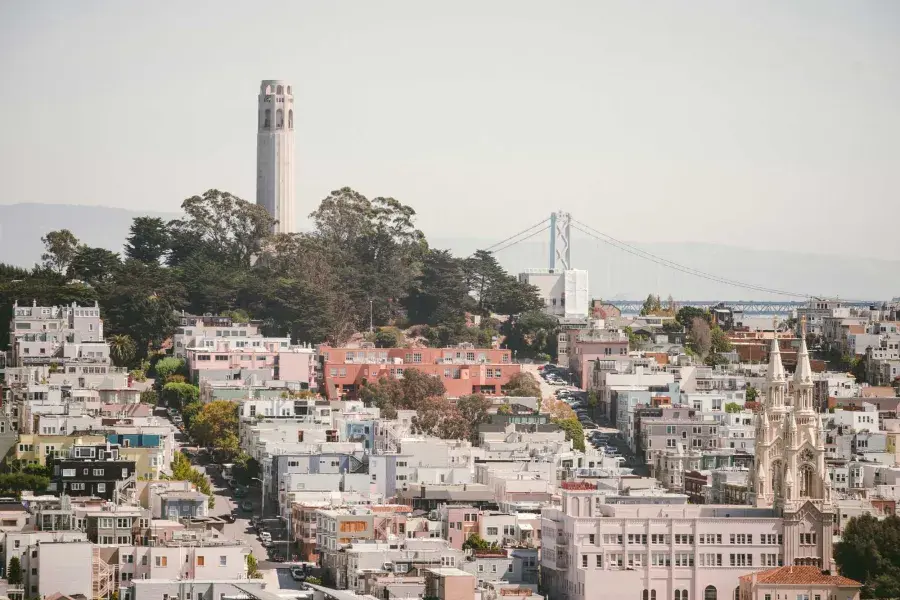 La Coit Tower de San Francisco est représentée avec le Bay Bridge en arrière-plan et une colline couverte de maisons au premier plan.
