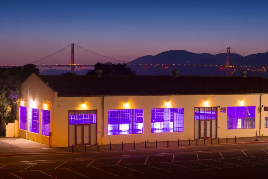 O edifício Fort Mason é iluminado por uma luz interna roxa à noite, com a ponte ao fundo.