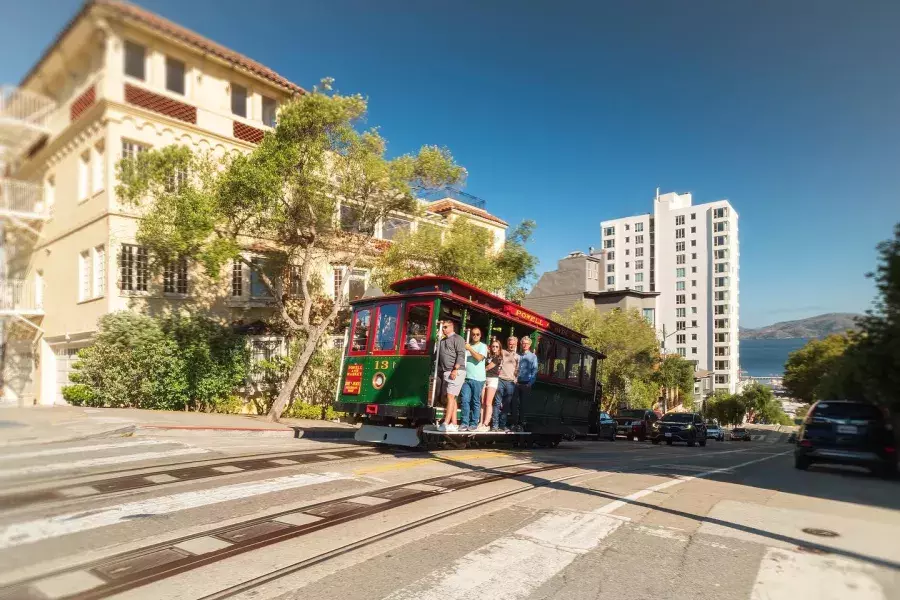 Powell and Hyde cable car going up the street with smiling people inside on a sunny day.