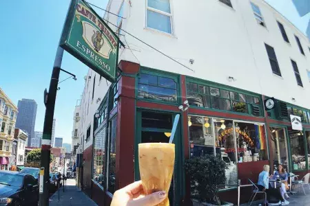 A person's hand holds a coffee drink with the exterior sign and storefront of Caffe Trieste in the background.