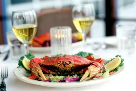 Um caranguejo Dungeness sentado em um prato em um restaurante com duas taças de vinho branco ao fundo.