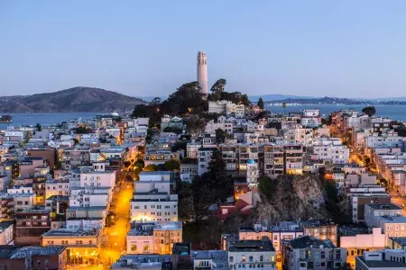 San Francisco's Coit Tower at dusk, with lighted streets before it and the San Francisco Bay behind it.