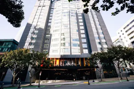 The exterior of the Clancy Hotel SF, tall grey and white building with an trees surrounding the front.