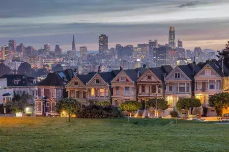 San Francisco's Painted Ladies, a row of Victorian houses, are pictured against a backdrop of the city skyline at twilight.