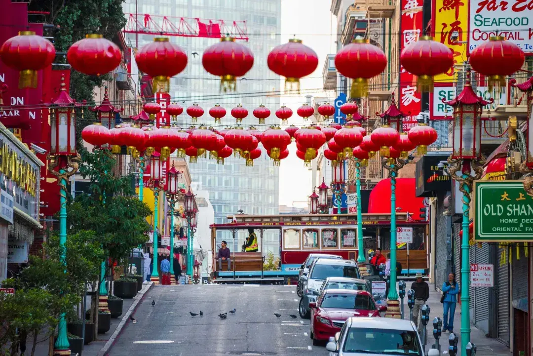 A hilly street in San Francisco's Chinatown is pictured with red lanterns dangling and a streetcar passing by.