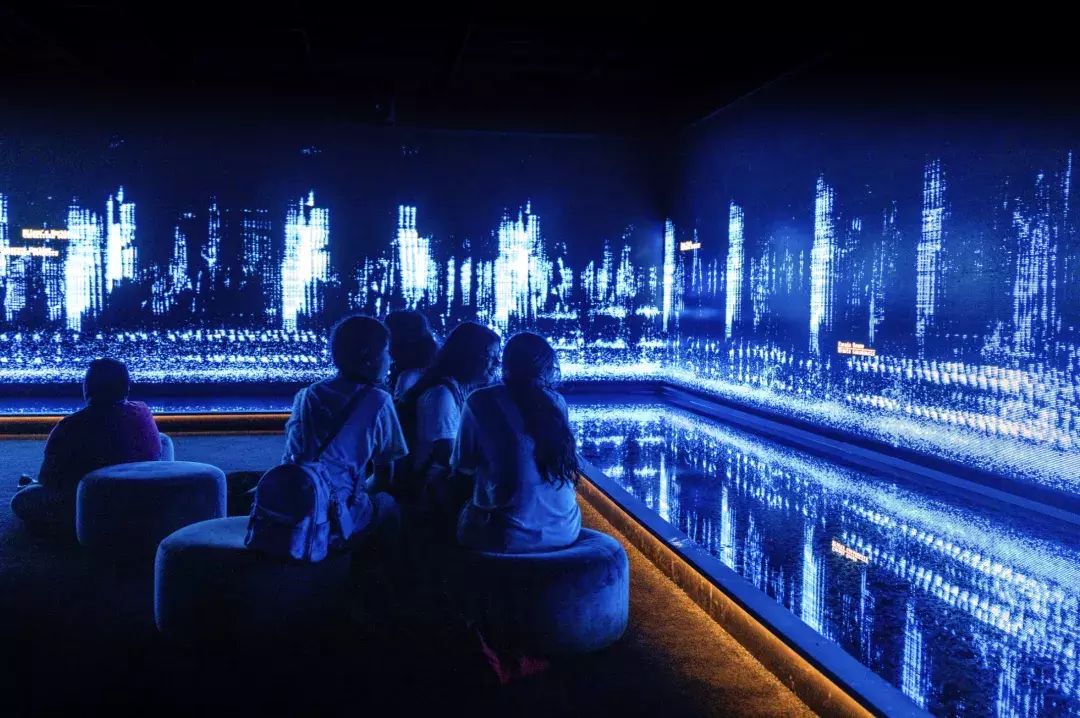 Children sit in a dark room, and look at an art installation creative of tiny moving lights on a wall