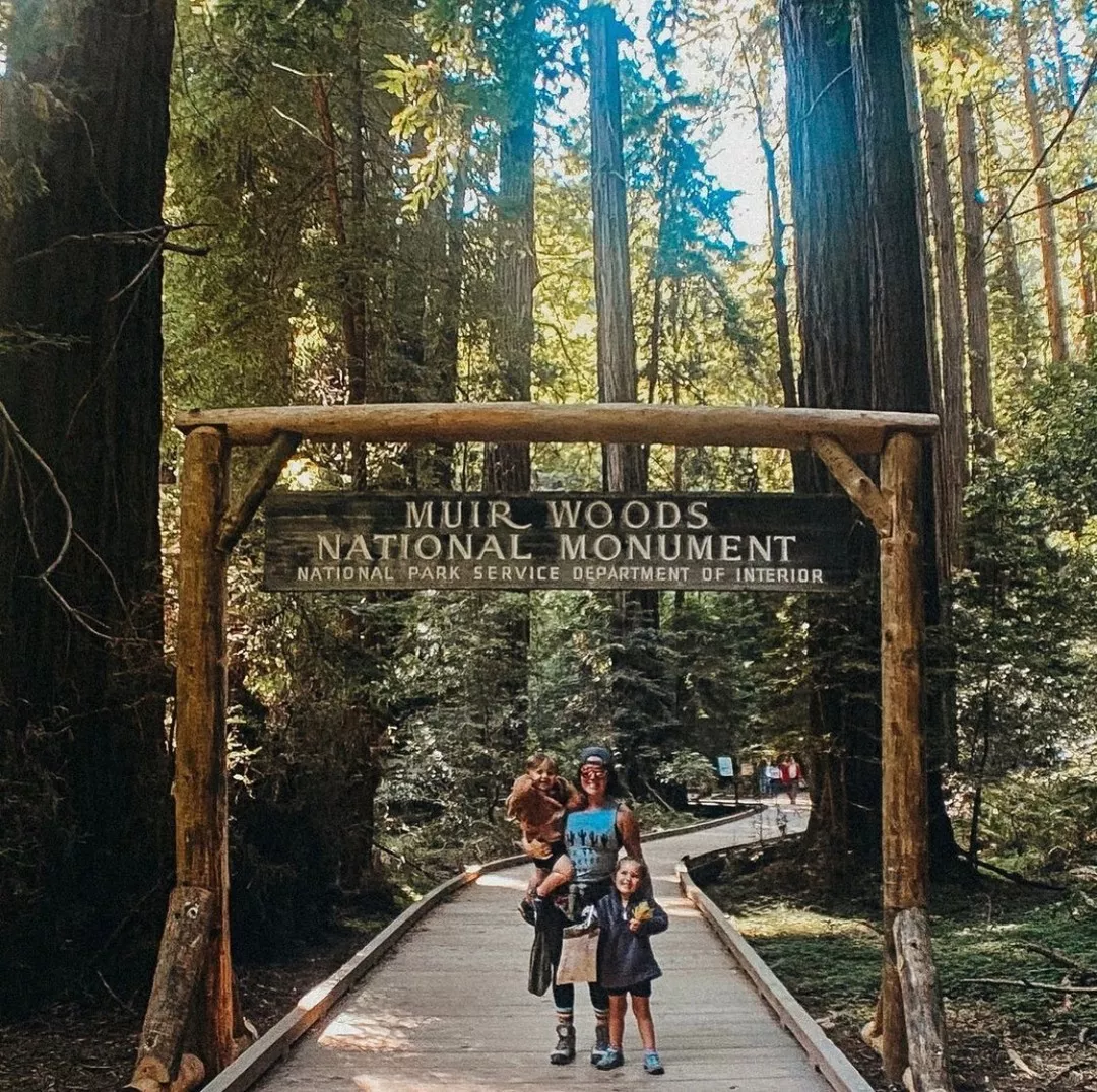 Woman and children stand in front of Muir Woods National Monument signage on a wooden trail in a forest