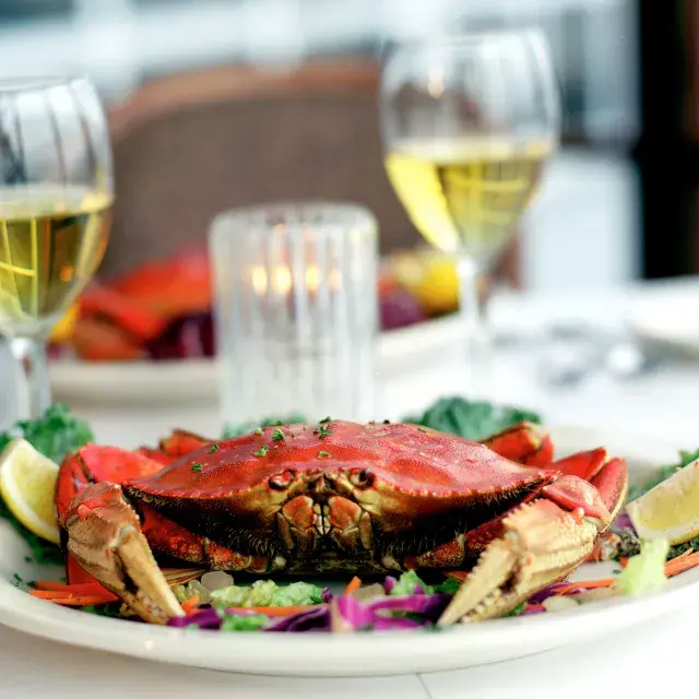 Um caranguejo Dungeness sentado em um prato em um restaurante com duas taças de vinho branco ao fundo.