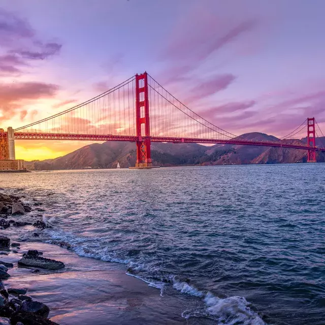 The Golden Gate Bridge at sunset with a multicolored sky and the San Francisco Bay in the foreground.