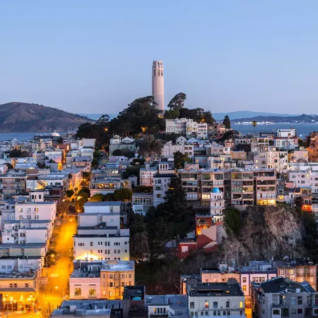 San Francisco's Coit Tower at dusk, with lighted streets before it and the San Francisco Bay behind it.