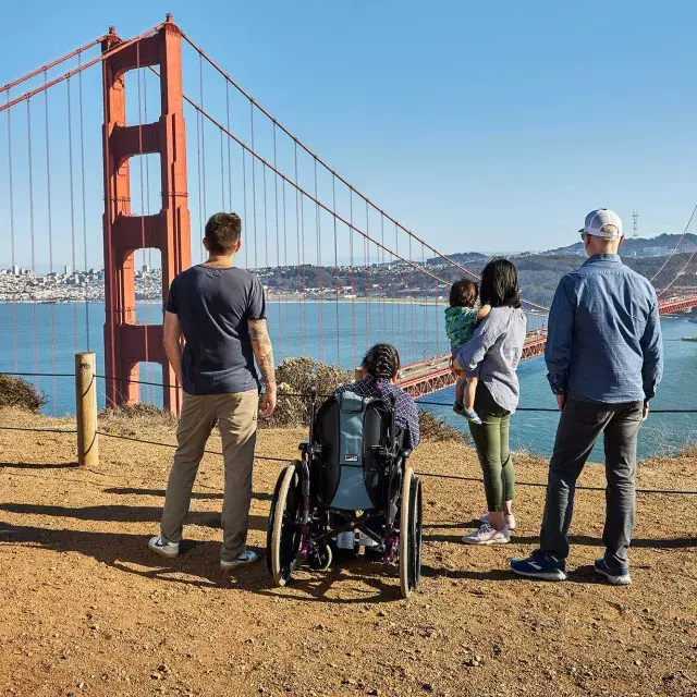 A group of people, including one person in a wheelchair, is seen from behind as they look at the Golden Gate Bridge from the Marin Headlands.