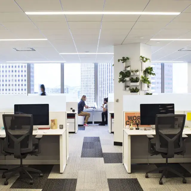 Interior scene at an office building in San Francisco, with desks in the foreground and two people working at a table in front of large windows.