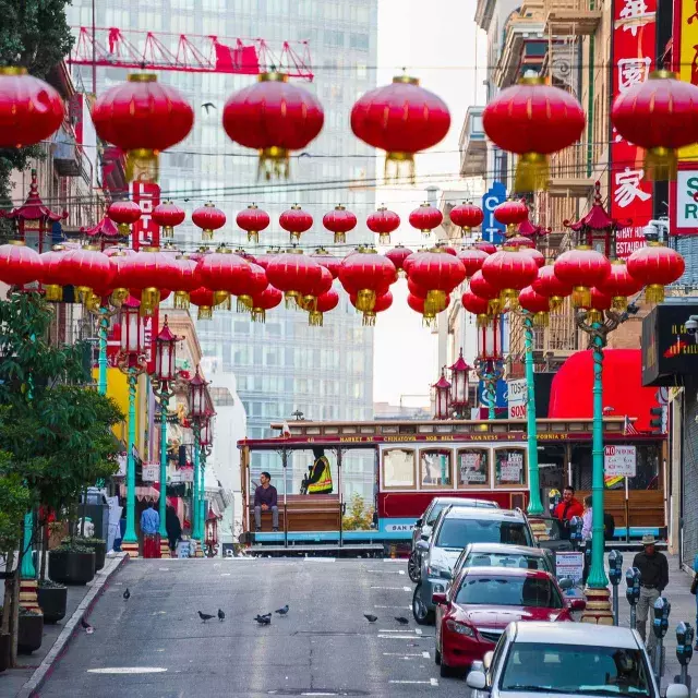 Uma rua montanhosa na Chinatown de São Francisco é retratada com lanternas vermelhas penduradas e um bonde passando.