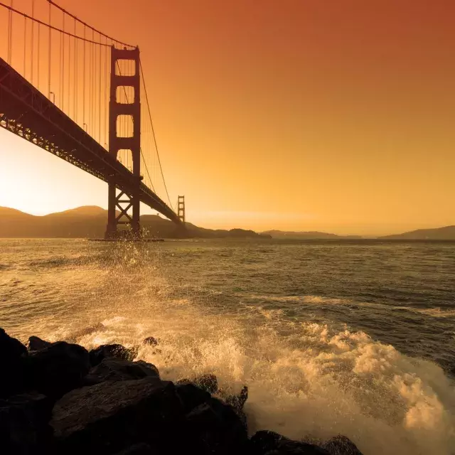 Waves crash near Fort Point beneath the Golden Gate Bridge at sunset.