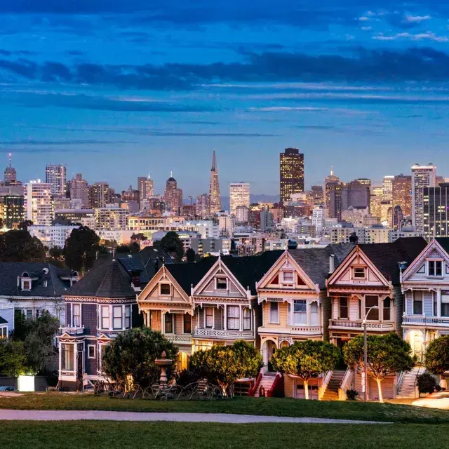 Die berühmten Painted Ladies vom Alamo Square sind in der Dämmerung vor der Skyline von San Francisco abgebildet.