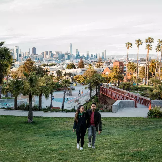 Ein Paar geht auf die Kamera zu, Dolores Park und die Skyline von San Francisco im Rücken.