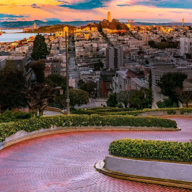Lombard Street curves with Coit Tower in the distance during sunset.