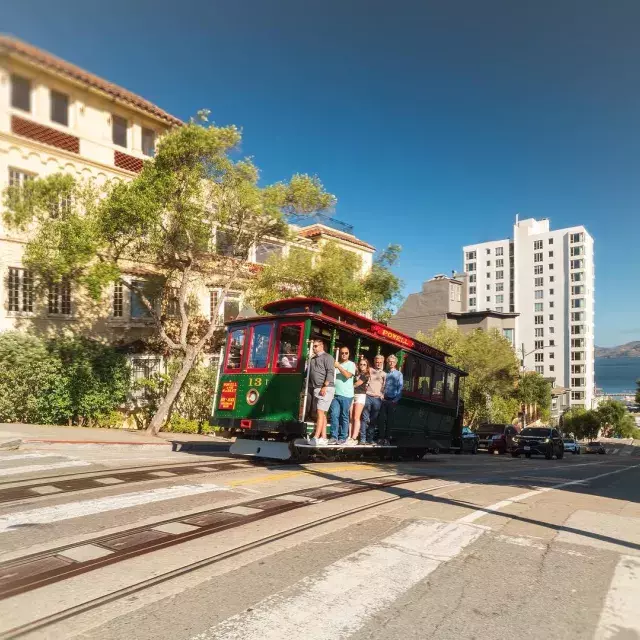 Powell and Hyde cable car going up the street with smiling people inside on a sunny day.