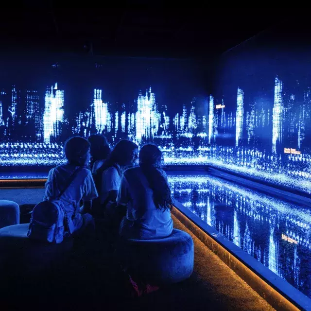 Children sit in a dark room, and look at an art installation creative of tiny moving lights on a wall