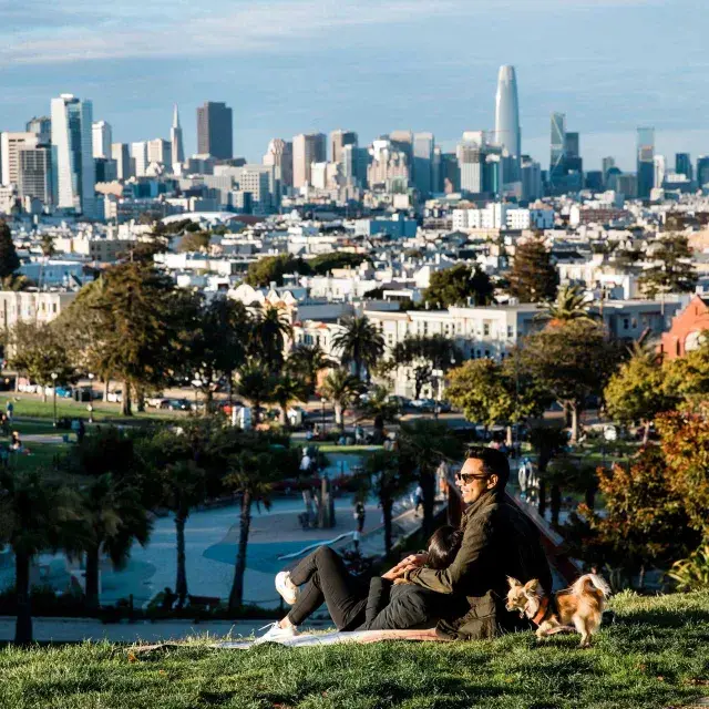 A man and woman sit in an embrace at Dolores Park while gazing at the San Francisco skyline. 